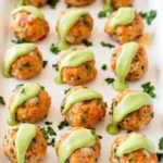 Baked salmon meatballs served on a white plate with a drizzle of creamy avocado sauce, garnished with fresh herbs.