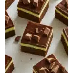 Freshly baked mint chocolate brownies stacked on a white plate, topped with a glossy chocolate ganache and garnished with mint leaves.